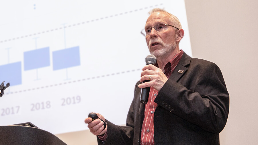 Speaker presenting Institutional Research information to a large crowd with a microphone and a powerpoint presentation.