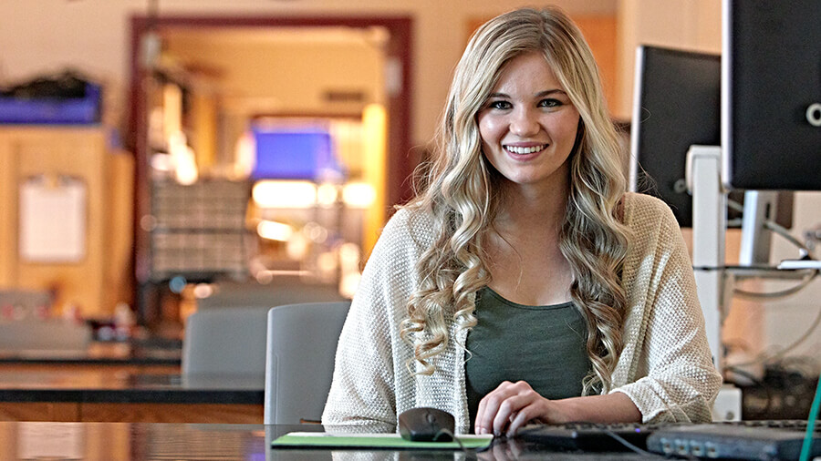 A student smiles for the camera while sitting at a computer.