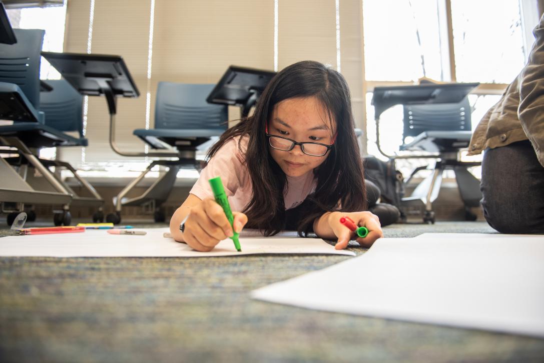 A student works on a large paper project on the floor.