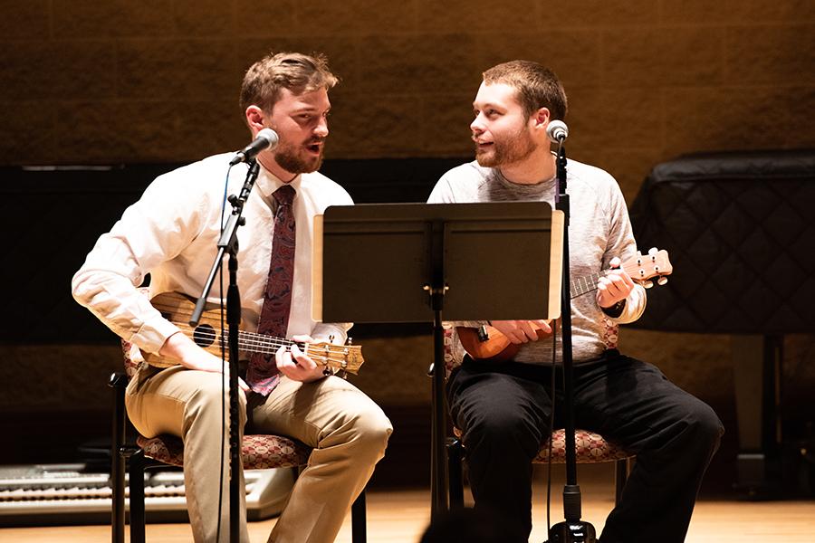 Two people sit with instruments in front of microphones.