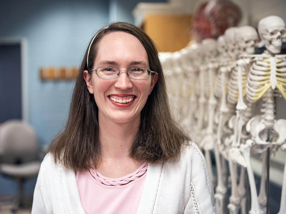 Regina Trout is standing in front of a row of human skeletons in a biology lab