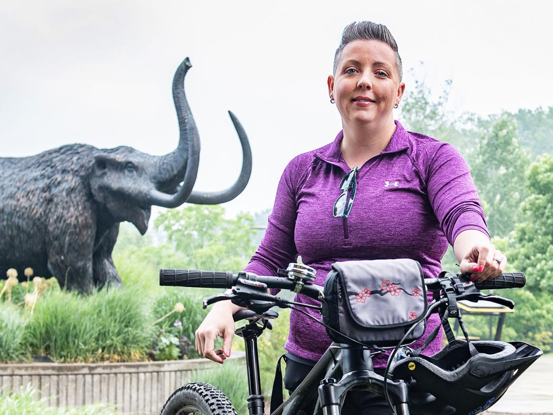 Teri Luce is standing in front of the mastodon statue with her bike.