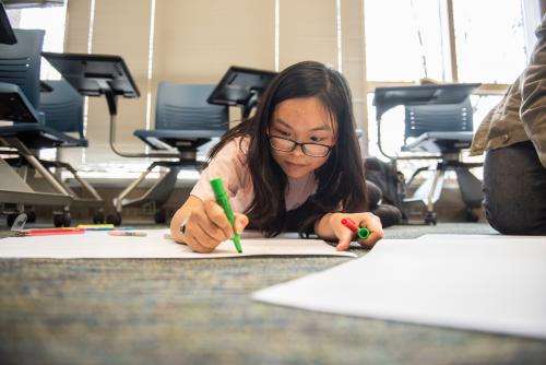 A student works on a large paper project on the floor.