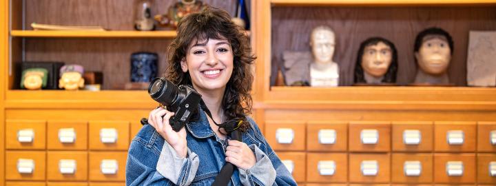 Bella Ferrin poses with a camera in an anthropology classroom.