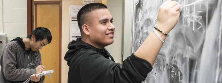 A student does math on a chalkboard while another student writes in a notebook.