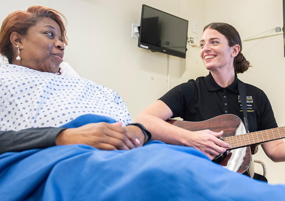 Music therapy student Charlene Delaney is playing the guitar and singing with a patient in a hospital bed.