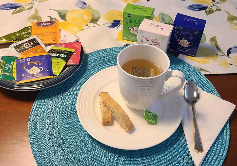 White mug with tea on top of white saucer with biscotti on a blue placemat with a plate a variety of tea packages in the background