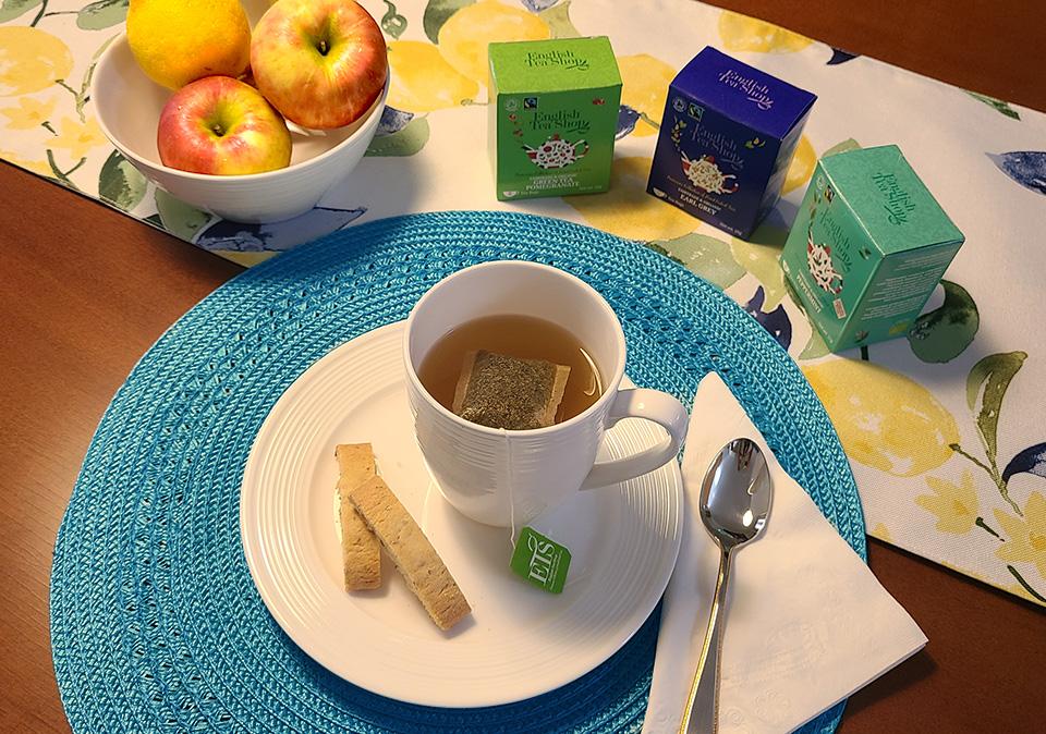 Tea in a white mug and biscotti on a white plate under a blue placemat on a table wth bowl of fruit and boxes of tea on a table runner with lemon print