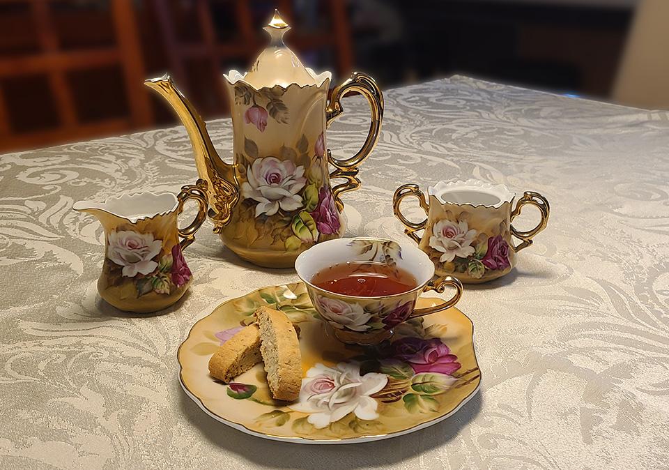 Antique tea set with a pink and white floral design on a fancy table cloth with a delicate cup full of tea on a matching plate with biscotti