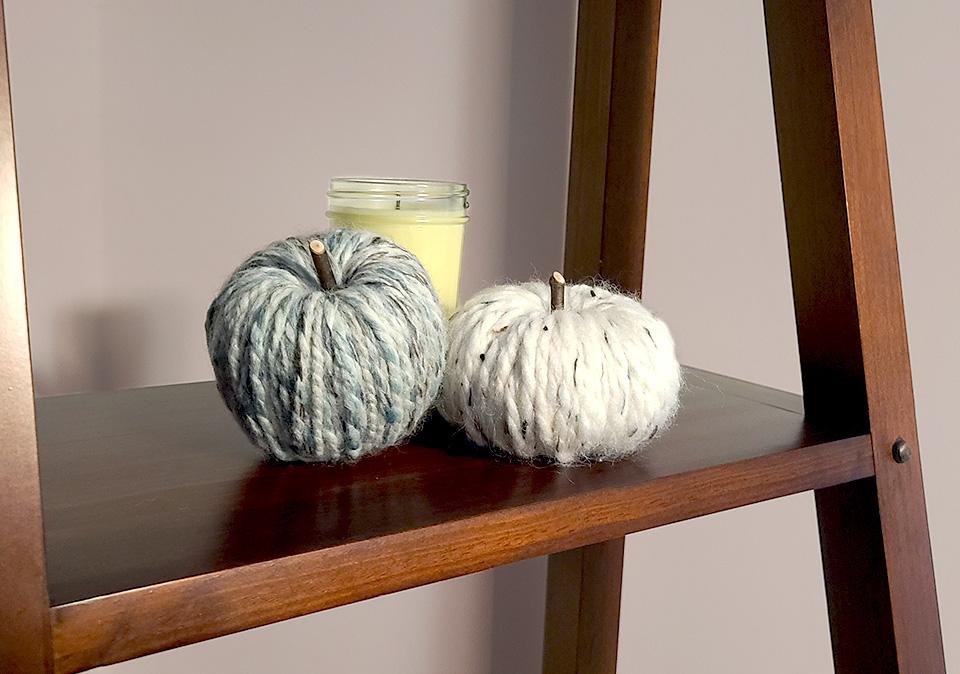 Two pumpkins made with white yarn and multi colr seafoam, gray, white yarn in front of a candle on a shelf stand.