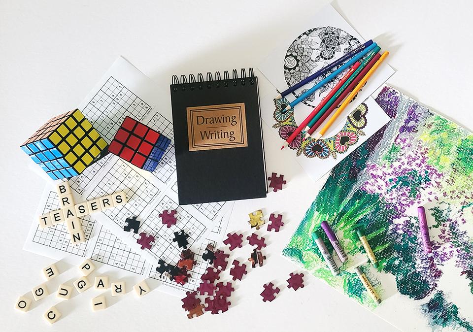 Photo montage of two Rubik's cubes, Sudoku, jisaw puzzle pieces, drawing/writing notebook, coloring book pages, colored pencils, and pastels on top of a pastel drawing.