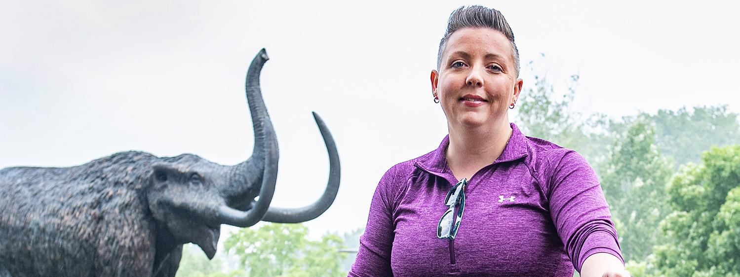 Teri Luce is standing in front of the mastodon statue with her bike.