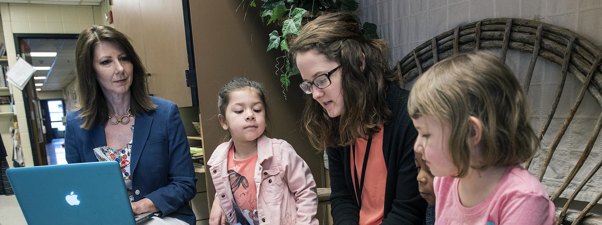 A female student teacher reads to children while faculty looks on