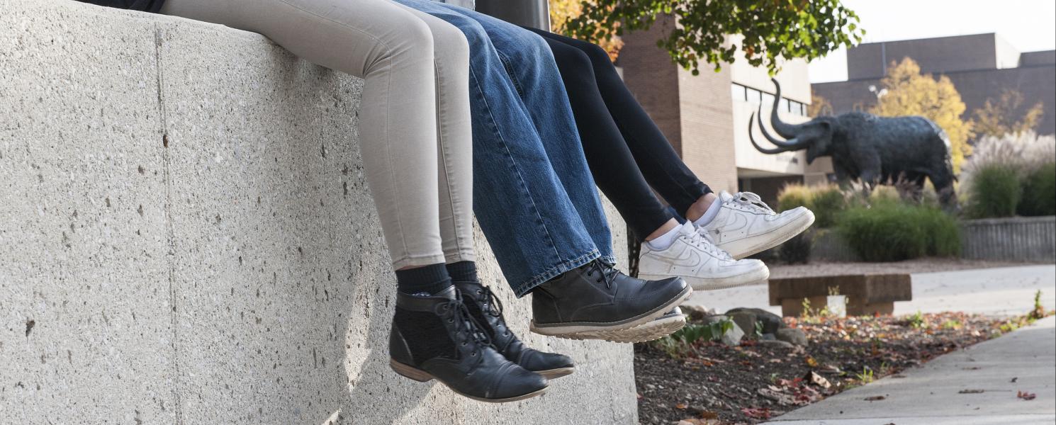 Close up photo of students' legs while they sit on a cement wall.