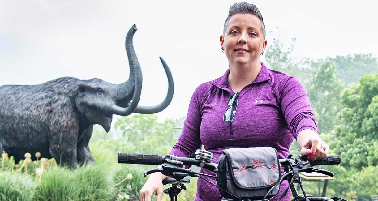 Teri Luce is standing in front of the mastodon statue with her bike.